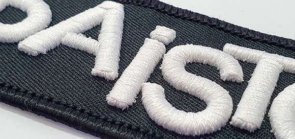 3D-Aufnäher Nahaufnahme: weiße gestickte 3D-Schrift auf einem schwarzen Grund.